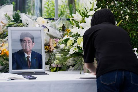 Eine Frau in schwarz beugt sich über einen Tisch mit weißer Decke, auf dem eine gerahmtes Foto eines alten Japaners steht