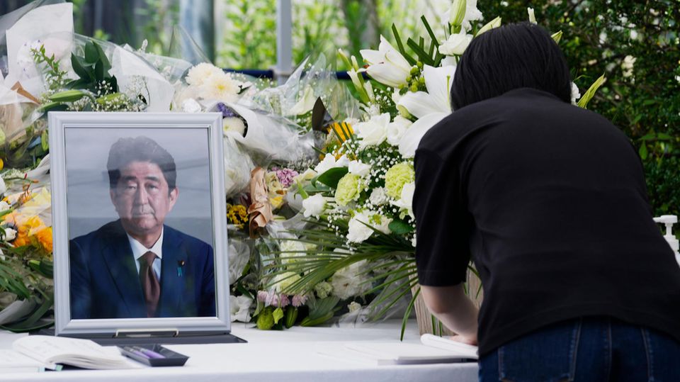 Eine Frau in schwarz beugt sich über einen Tisch mit weißer Decke, auf dem eine gerahmtes Foto eines alten Japaners steht