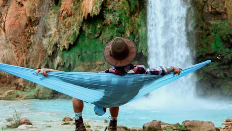 Mann mit Hut sitzt vor einem Wasserfall auf einer Hängematte.