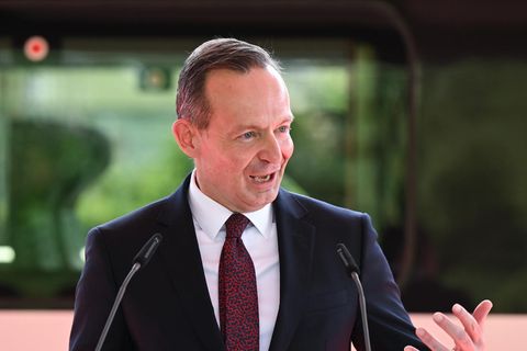 Ein weißer Mann im Anzug gestikuliert hinter einem weißen Rednerpult mit rotem "DB"-Logo. Hinter ihm ein ICE