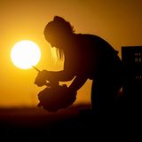 Im Gegenlicht der aufgehenden Sonne steht eine Frau auf einem Feld und hält ein Messer in der einen, Salat in der anderen Hand