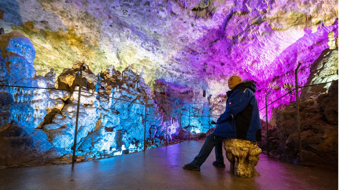 Ein ältere Mann in blau-schwarzem Annorak sitzt auf einem Stein mitten in einer bunt angeleuchteten Höhle