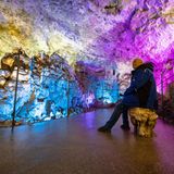 Ein ältere Mann in blau-schwarzem Annorak sitzt auf einem Stein mitten in einer bunt angeleuchteten Höhle