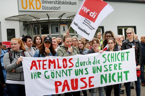 Frauen und Männer stehen mit einem Banner "Atemlos durch unsere Schicht, Ihr gönnt uns unsere Pause nicht vor einem Krankenhaus