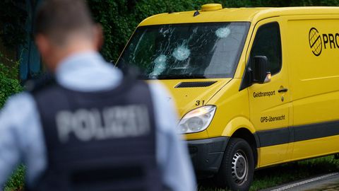 Ein weißer gepanzerter Transporter steht auf dem Seitenstreifen einer mehrspurigen Straße. Am Heck stehen Polizisten in Uniform