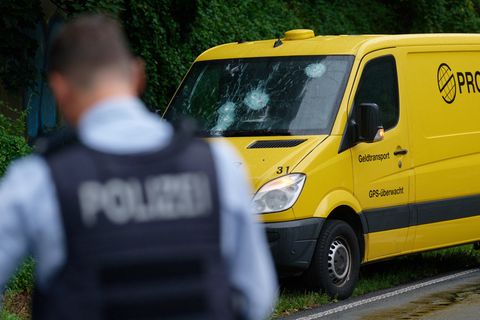 Ein weißer gepanzerter Transporter steht auf dem Seitenstreifen einer mehrspurigen Straße. Am Heck stehen Polizisten in Uniform