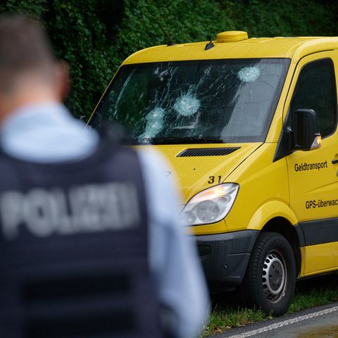 Ein weißer gepanzerter Transporter steht auf dem Seitenstreifen einer mehrspurigen Straße. Am Heck stehen Polizisten in Uniform