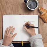 Eine Frau schreibt in ihre Tagebuch an einem Tisch mit Kaffee und Croissant.