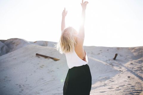 Eine Frau streckt bei Sonnenschein am Strand ihre Hände in die Luft.