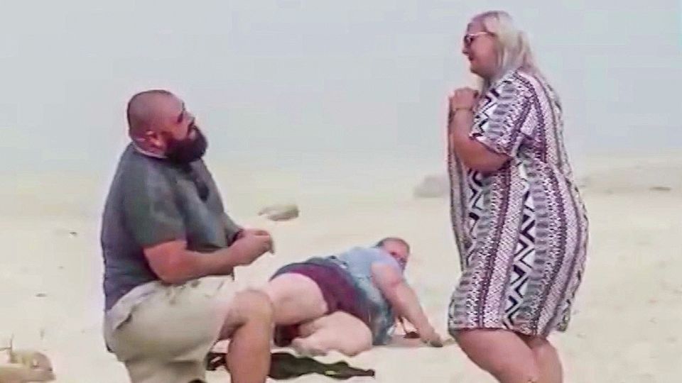 Ein Mann macht einer Frau einen Heiratsantrag am Strand – ihre Schwester stürzt im Hintergrund.