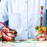 Mann sitzt mit Farbe an den Händen und Pinseln in der Hand am Tisch.