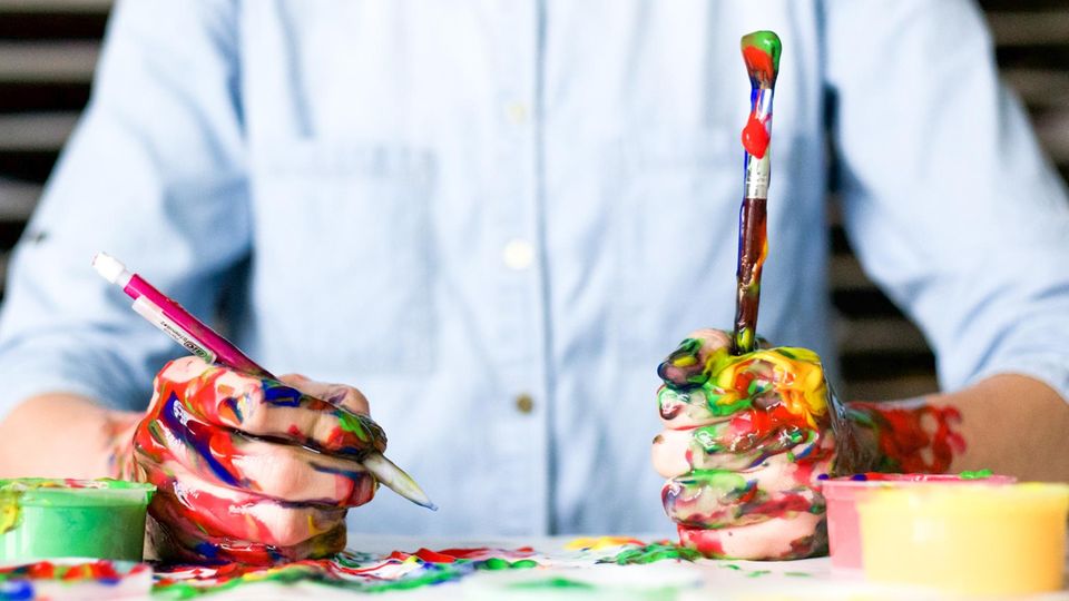 Mann sitzt mit Farbe an den Händen und Pinseln in der Hand am Tisch.