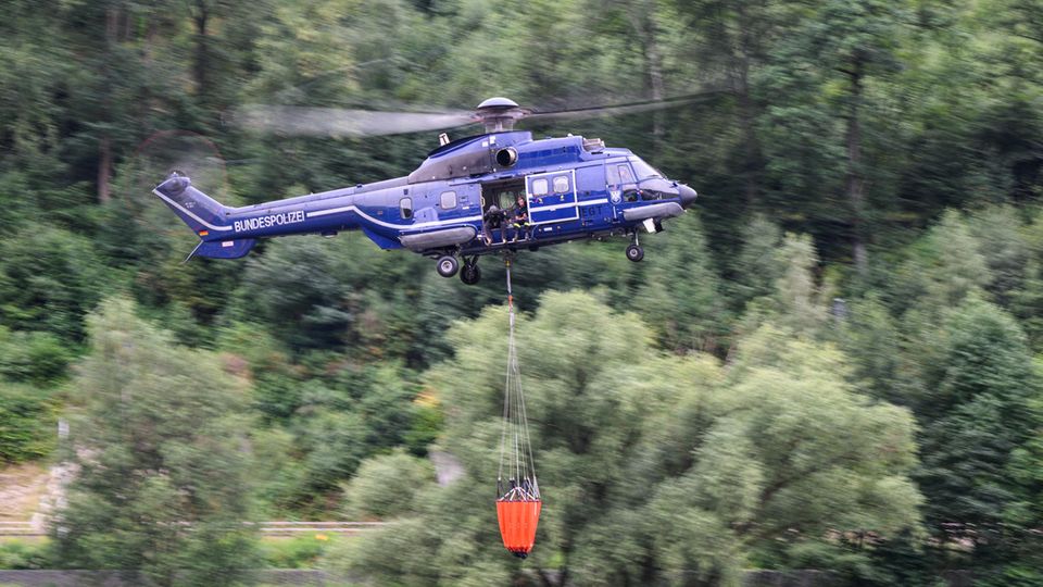 Ein blauer Hubschrauber mit Bundespolizei-Aufschrift fliegt mit orangem Löschsack unter dem Rumpf über einen Wald