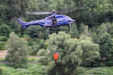 Ein blauer Hubschrauber mit Bundespolizei-Aufschrift fliegt mit orangem Löschsack unter dem Rumpf über einen Wald