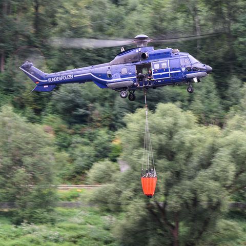 Ein blauer Hubschrauber mit Bundespolizei-Aufschrift fliegt mit orangem Löschsack unter dem Rumpf über einen Wald
