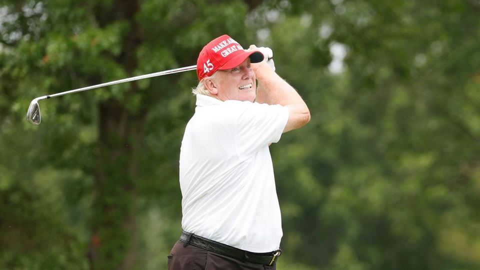 Donald Trump in weißem Hemd und roter Schirmmütze schwingt den Golfschläger beim Abschlag