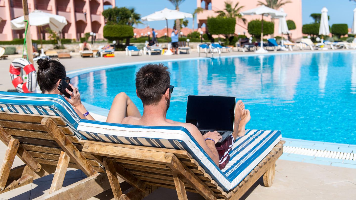 Ein Mann und eine Frau sind von hinten zu sehen, wie sie im Urlaub mit Laptop und Handy auf Liegen am Pool liegen