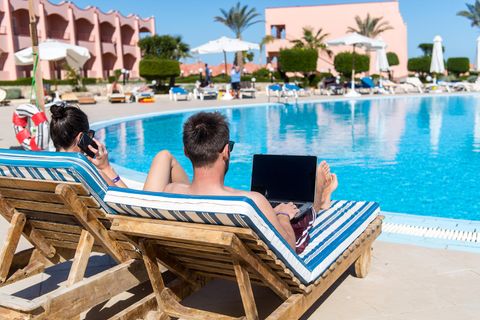 Ein Mann und eine Frau sind von hinten zu sehen, wie sie im Urlaub mit Laptop und Handy auf Liegen am Pool liegen