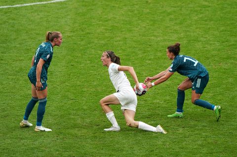 Eine weiße Fußballspielerin ganz in Weiß springt mit weit aufgerissenem Mund auf und nähert sich ihrer Gegenspielerin in Grün