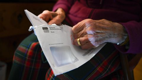 Faltige Frauenhände ziehen einen Rentenbescheid aus einem grauen, länglichen Briefumschlag der Deutschen Rentenversicherung