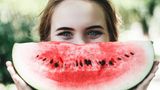Eine Frau grinst mit einer Wassermelone vor dem Mund in die Kamera.