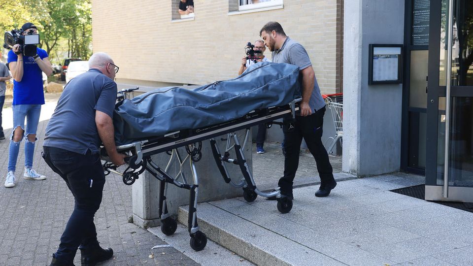 Zwei weiße Männer in schwarzen Hosen und blauen Polohemden schieben eine Bahre mit Leichensack aus einem Wohnhaus
