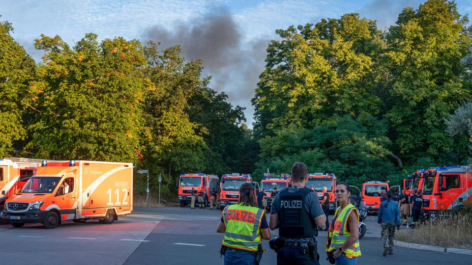 Polizeibeamte und Feuerwehrfahrzeuge stehen am Kronprinzessinnenweg in Berlin. Im Berliner Grunewald ist nach einer unbeabsichtigten Explosion auf dem dortigen Sprengplatz am Donnerstagmorgen ein Feuer ausgebrochen und hat den angrenzenden Wald in Brand gesetzt. 