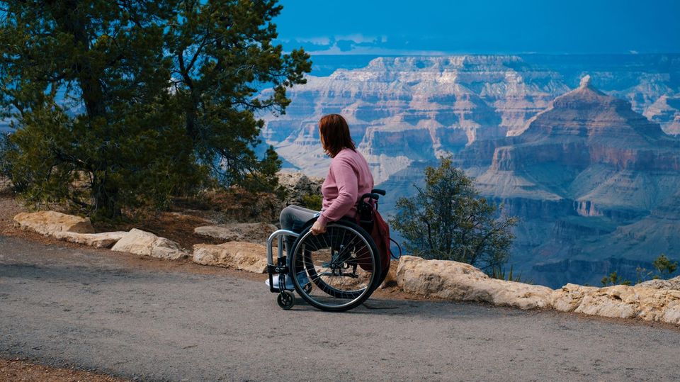 Eine Frau im Rollstuhl blickt auf eine Berglandschaft.