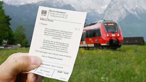 Ein Mann hält ein 9-Euro-Ticket in der Hand, im Hintergrund fährt ein Zug der Deutschen Bahn