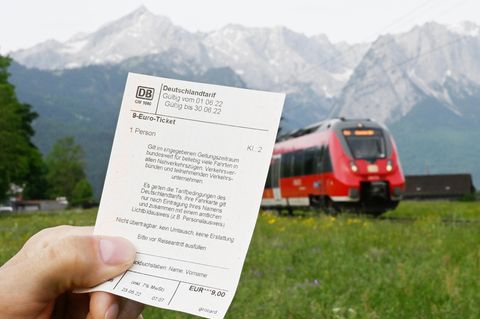 Ein Mann hält ein 9-Euro-Ticket in der Hand, im Hintergrund fährt ein Zug der Deutschen Bahn