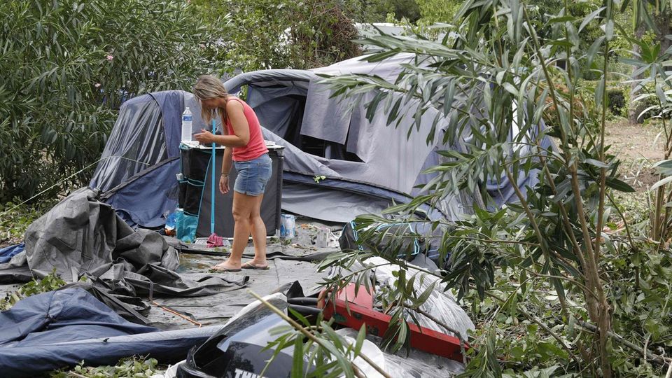 Eine weißte Frau in rotem Top und blauen Shorts steht auf einem Zeltplatz zwischen umgestürzten Bäumen und zerstörten Zelten