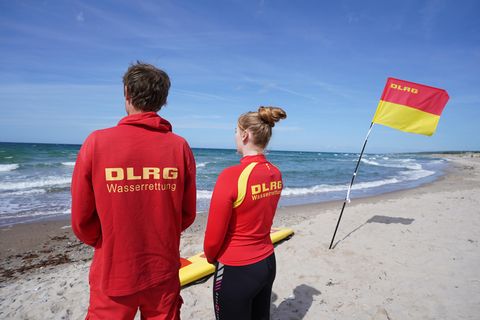 Ein junger Mann und eine junge Frau in roter Kleidung mit gelber "DLRG"-Schrift schauen an gespannt auf die sonnige See hinaus