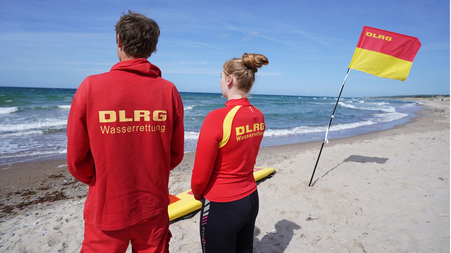 Ein junger Mann und eine junge Frau in roter Kleidung mit gelber "DLRG"-Schrift schauen an gespannt auf die sonnige See hinaus