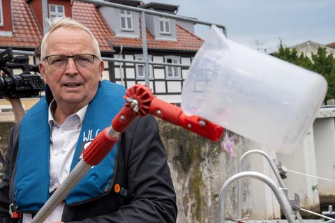 Ein älterer weißer Mann mit Brille, weißem Seitenscheitel und blauem Anzug hält eine Stange mit einem Messbecher in die Kamera
