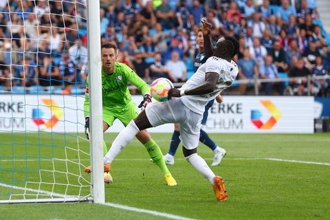 Ein schwarzer Fußball-Profi in weißem Trikot hat am linken Torpfosten den Ball an der linken Faust