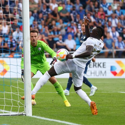 Ein schwarzer Fußball-Profi in weißem Trikot hat am linken Torpfosten den Ball an der linken Faust