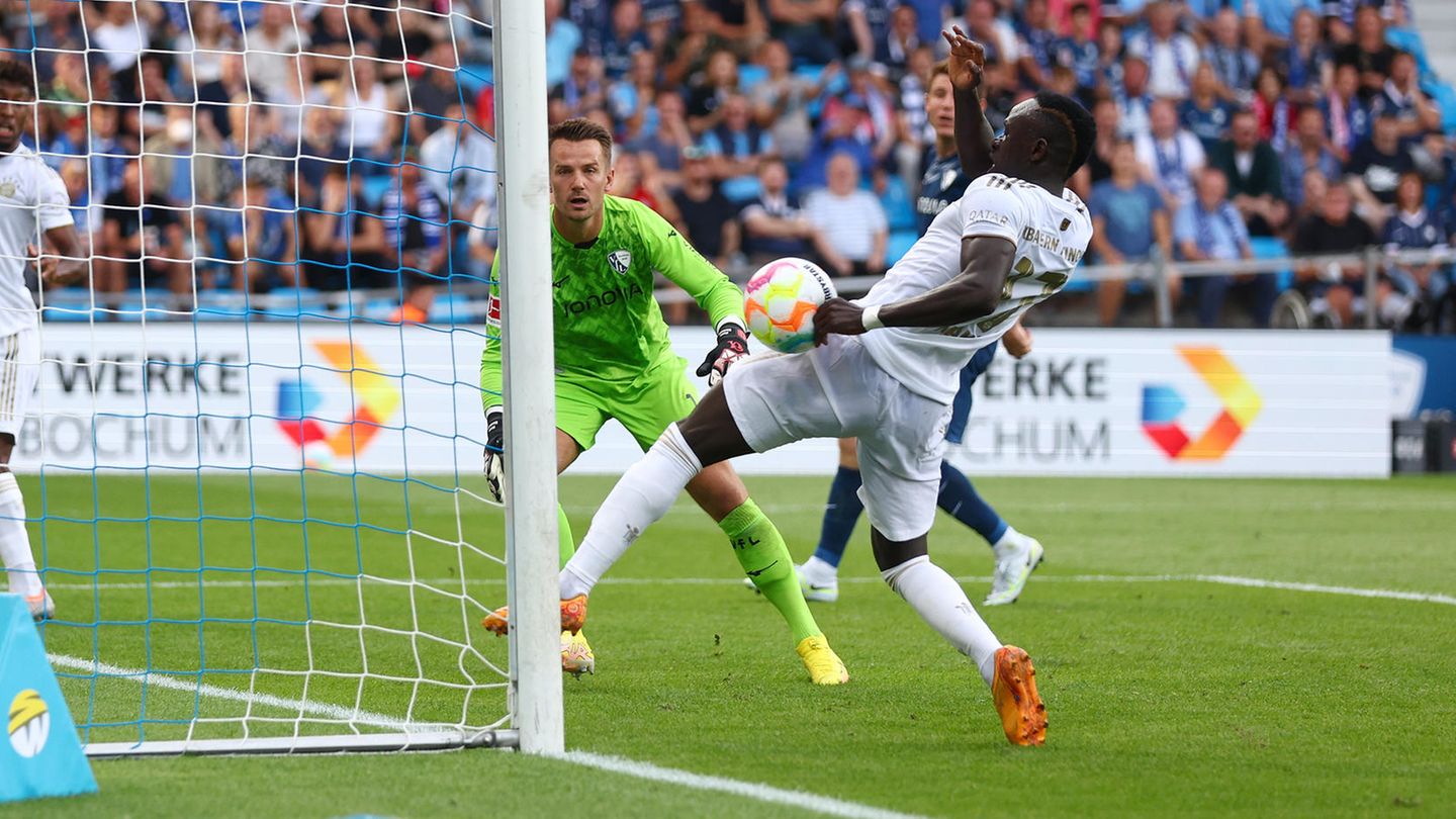 Ein schwarzer Fußball-Profi in weißem Trikot hat am linken Torpfosten den Ball an der linken Faust