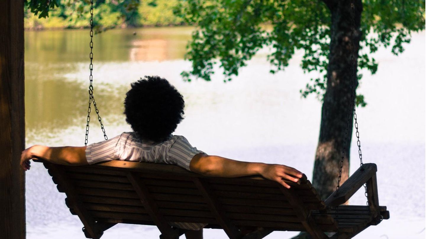 Ein Mann sitzt in einer Hollywoodschaukel und blickt auf einen See.