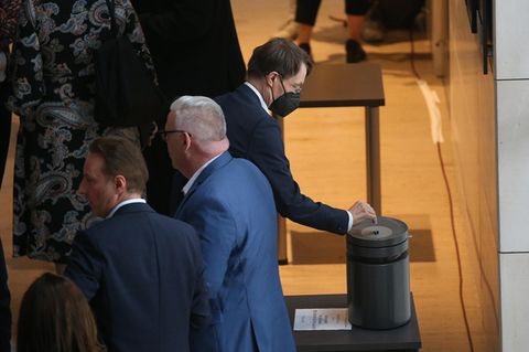 Ein Mann im Anzug steckt eine Stimmkarte in ein grauen Metallzylinder mit einem Schlitz oben