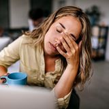 Eine Frau sitzt mit einem Kaffee am Tisch und fasst sich gestresst an den Kopf.