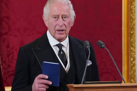 Charles III. hält in einer Hand ein Buch und steht vor zwei Mikrofonen