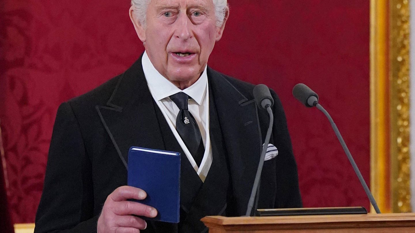 Charles III  holds a book in one hand and stands in front of two microphones