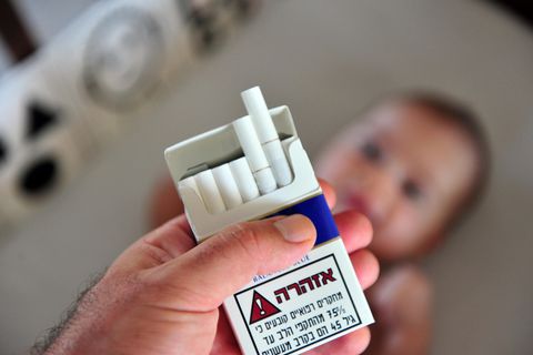 Eine männliche Hand hält eine Zigarettenschachtel, im Hintergrund liegt ein Baby (Symbolbild Passivrauchen)