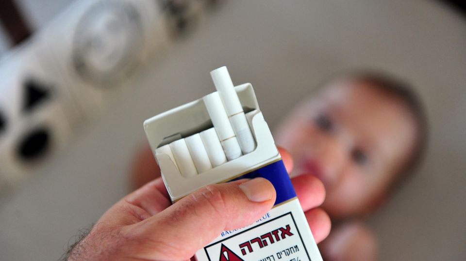 Eine männliche Hand hält eine Zigarettenschachtel, im Hintergrund liegt ein Baby (Symbolbild Passivrauchen)