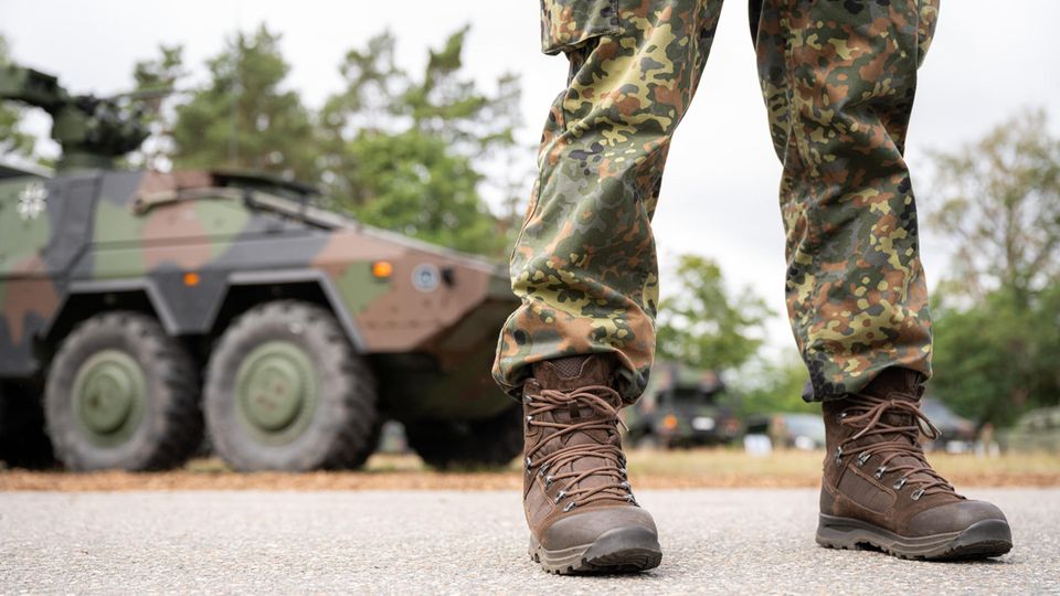 Beine und Stiefel eines Bundeswehr-Soldaten in Tarnfleck-Uniform stehen vor einem Truppentransporter in Tarnlackierung