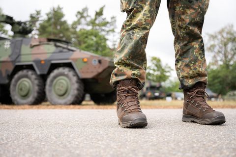 Beine und Stiefel eines Bundeswehr-Soldaten in Tarnfleck-Uniform stehen vor einem Truppentransporter in Tarnlackierung