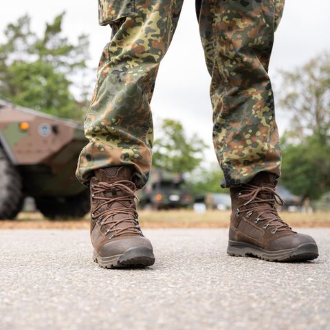 Beine und Stiefel eines Bundeswehr-Soldaten in Tarnfleck-Uniform stehen vor einem Truppentransporter in Tarnlackierung