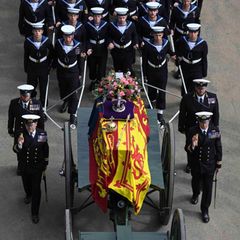 Auf einer Laffette liegt ein mit einer rot-gelben Flagge verdeckter Sarg. Soldaten in Ehrenuniform begleiten ihn