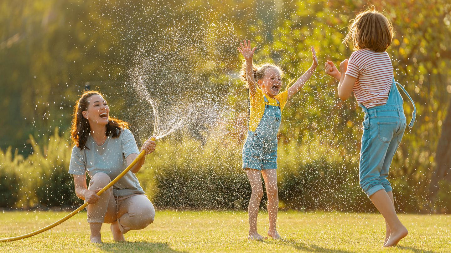 Eine Frau spritzt mit einem Wasserschlauch zwei Mädchen nass.