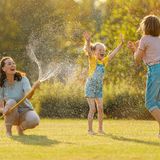 Eine Frau spritzt mit einem Wasserschlauch zwei Mädchen nass.